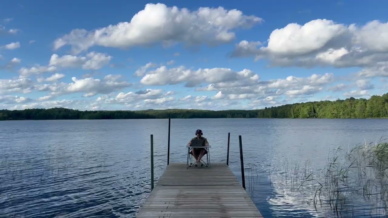 A lone figure seated at the end of a wooden jetty on a calm lake, forest along the far shore under a bright blue sky